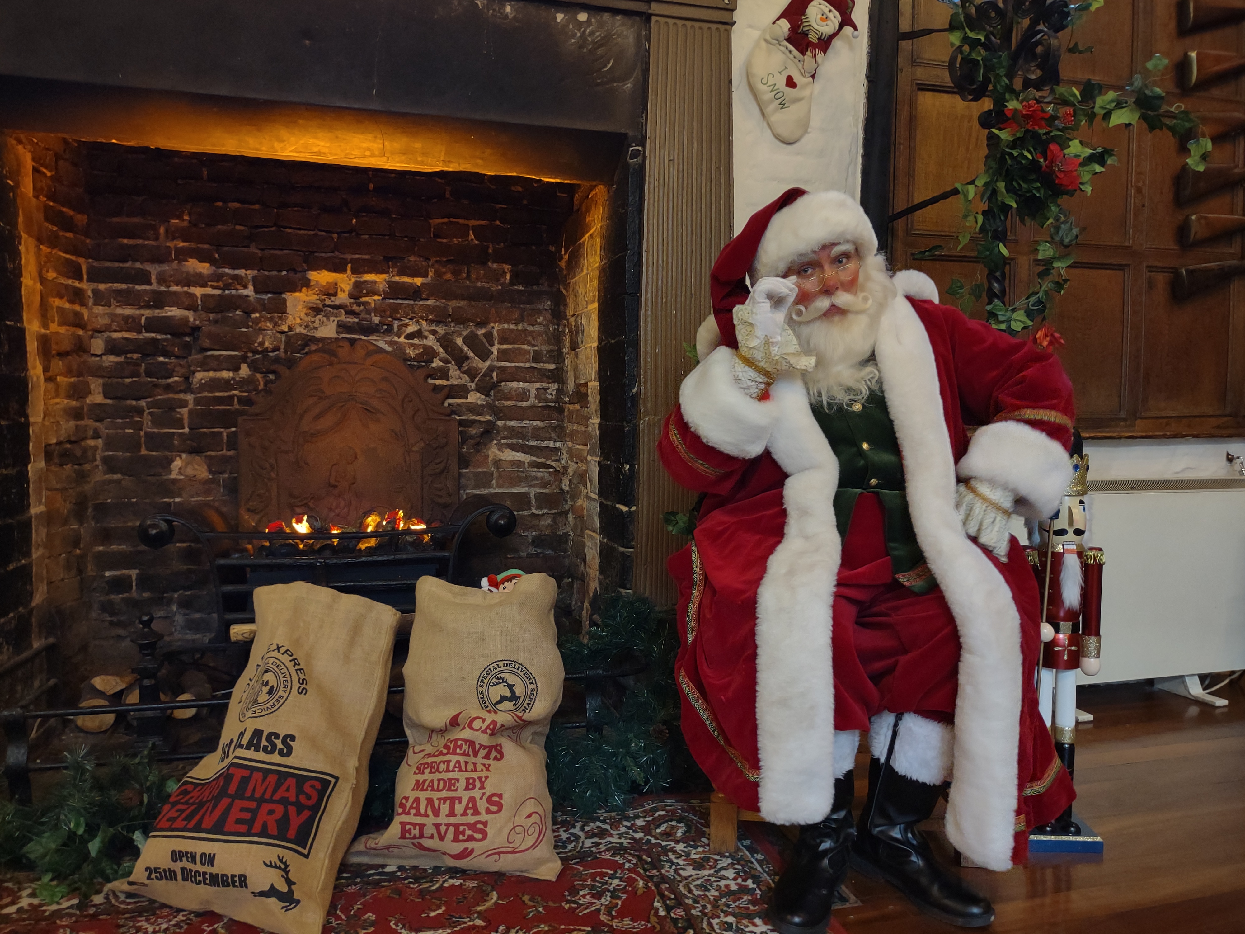 A man dressed as Father Christmas, in red and white attire with a white beard and glasses. Sat on a chair next to a fireplace