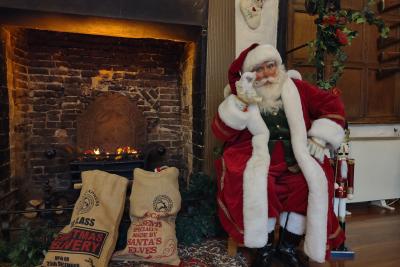 A man dressed as Father Christmas, in red and white attire with a white beard and glasses. Sat on a chair next to a fireplace